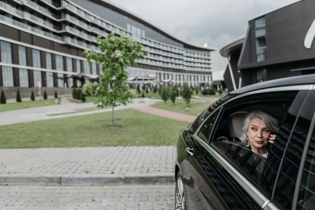 Elderly woman with grey hair on a phone call sitting in car near modern building.